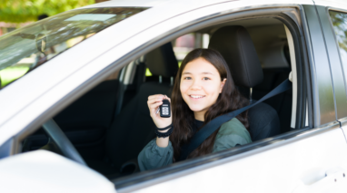 Smiling female teenager sitting on the drivers seat of a car holding up the keys