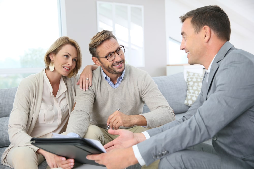 A smiling insurance agent explains a car insurance policy to a couple sitting on a couch.