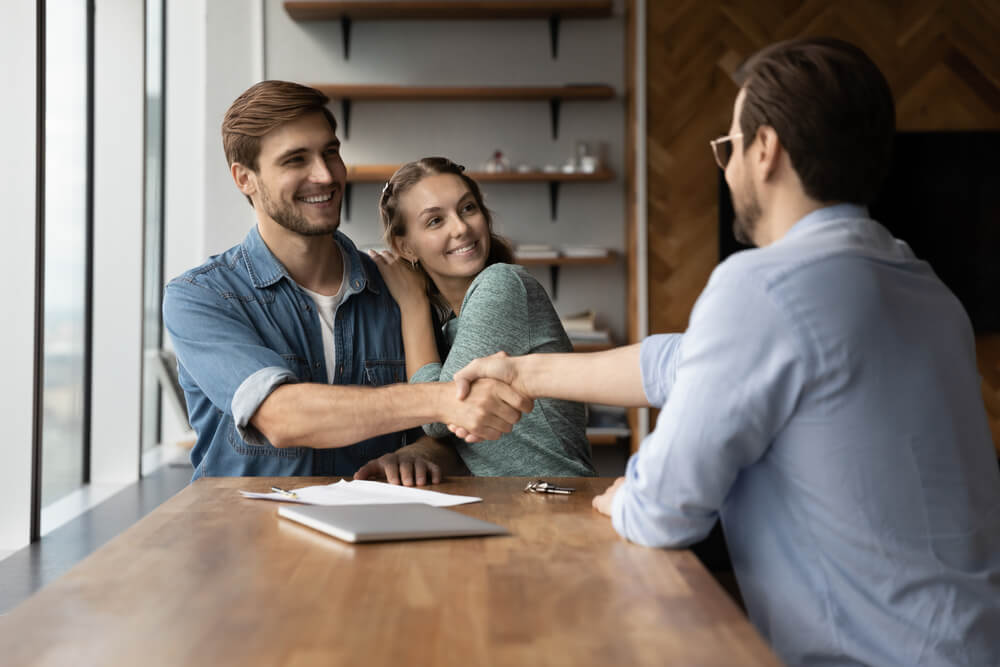 A new insurance agent in the Mid-Atlantic region seen from behind shakes hands with a young couple who are happy clients.