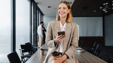 Smiling woman insurance agent sits on board room table with phone in hand.