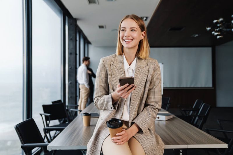 Smiling woman insurance agent sits on board room table with phone in hand.